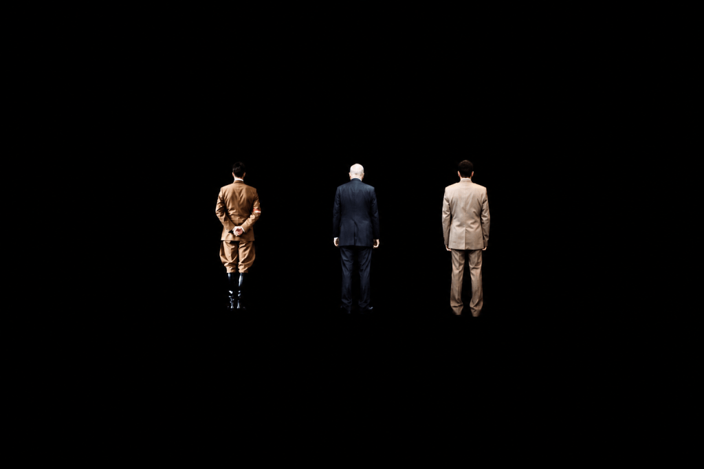 Three men standing with their backs to the viewer against a dark background; one in a military uniform, one in a suit, and one in a light suit.