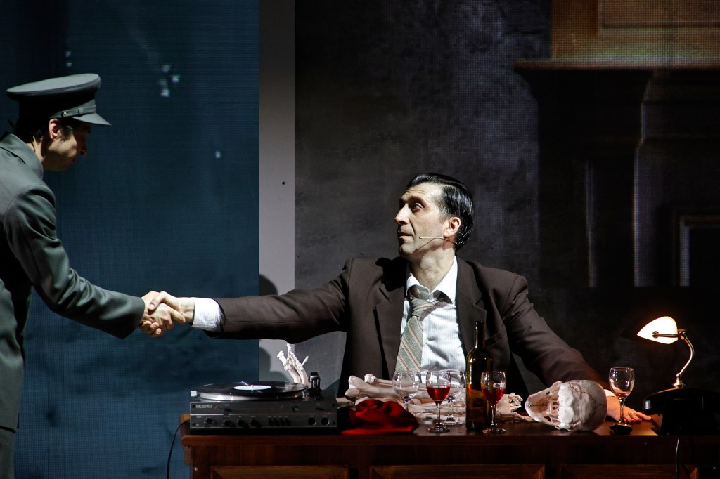 A dramatic scene with two men shaking hands; one is in military attire while the other is dressed in a brown suit, seated at a desk with a record player, drinks, and a skull.