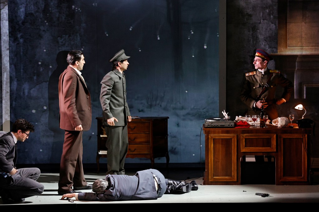 A dramatic stage scene featuring four male actors, with one lying on the floor and the others standing. The characters are dressed in period costumes, with a desk and props in the background, set against a dark, atmospheric backdrop.
