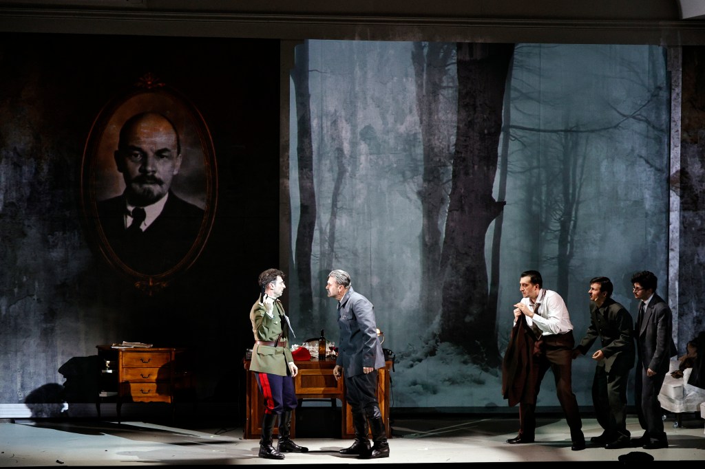 A dramatic scene from a theatrical performance featuring two characters in confrontation, set against a backdrop of a forest and a large portrait of Lenin. Several other characters are present, observing the exchange.