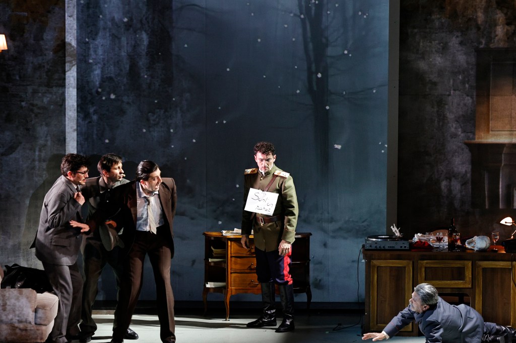 A dramatic theatrical scene featuring a group of men in suits confronting a military officer holding a sign. The background depicts a snowy landscape, enhancing the tense atmosphere.