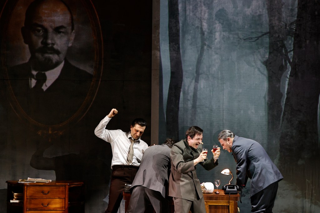 A theatrical scene featuring several men in period costumes, celebrating or toasting with drinks, against a backdrop that includes a portrait of a historical figure and a forest landscape.