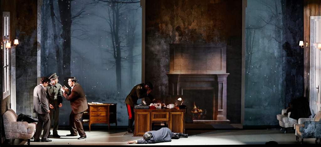 A dramatic stage scene featuring several actors in period costumes engaged in intense conversation, with a winter landscape backdrop and a fireplace set piece.