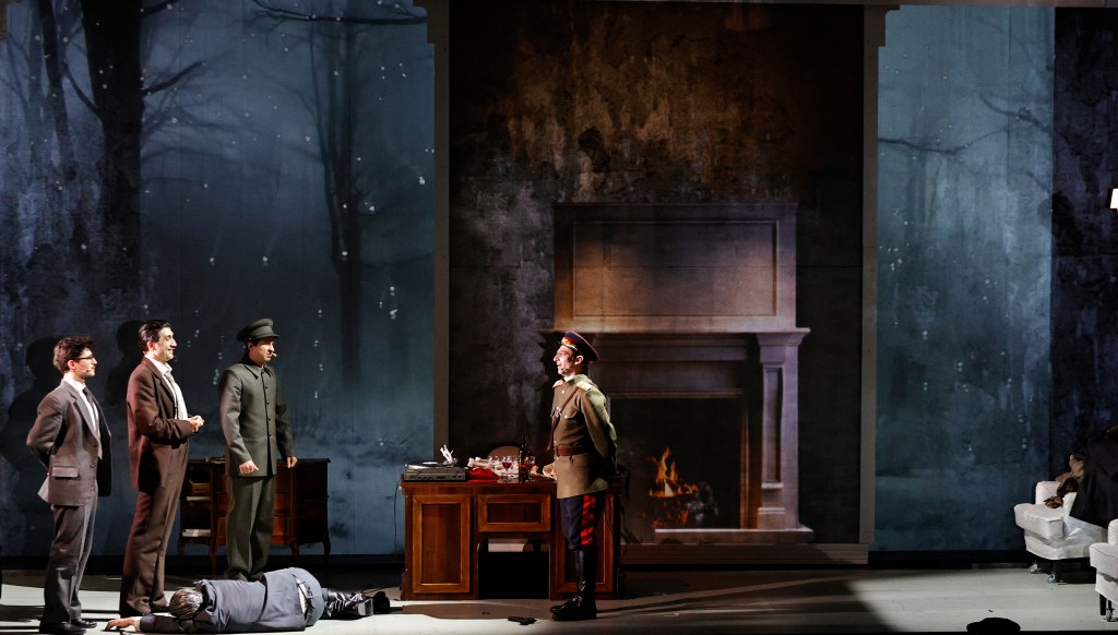 A theatrical scene featuring four actors in formal attire, standing on a stage with a backdrop of a dark, forested landscape and an interior setting that includes a fireplace. One actor lies on the ground in a dramatic pose, while another, dressed in a military uniform, appears to be addressing the other characters.
