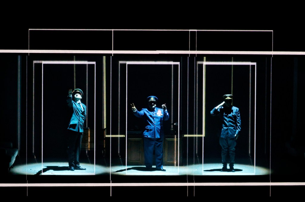 Three actors dressed in military uniforms performing on stage, with dramatic lighting and a dark background.