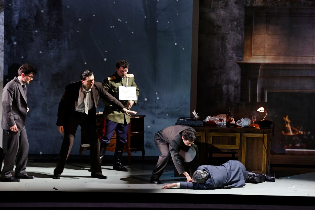 A dramatic scene from a theatrical performance featuring five male actors. One actor in a military uniform holds a sign, while others display various expressions of concern and urgency as one man lies on the floor, suggesting a critical moment.