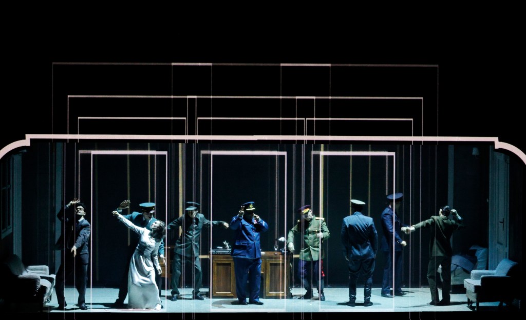 A theatrical performance featuring several actors in dramatic poses, set against a stylized backdrop resembling a room with glass partitions. The cast wears period costumes, with a mix of military and civilian attire, creating a tense atmosphere.