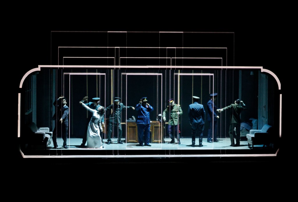 A theatrical performance featuring a group of actors in period costumes, including a woman in a white dress and men in military uniforms, staged in a stylized set with multiple layered frames. The actors are expressing various poses and emotions, suggesting a dramatic or tense scene.