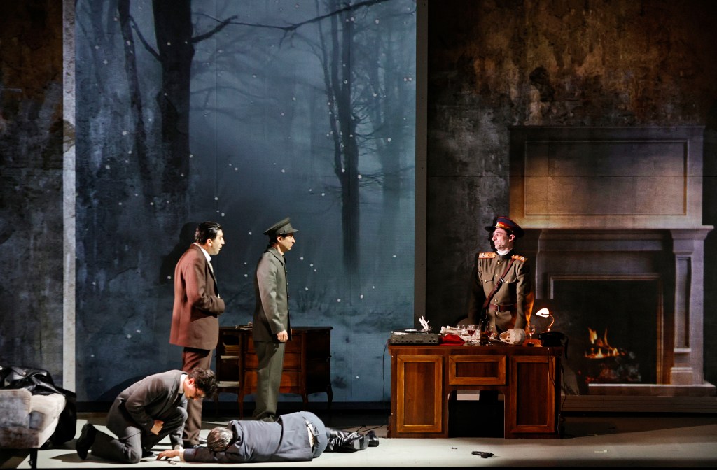 A scene from a theatrical performance set in a dimly lit room with a fireplace, featuring four characters: two standing, one kneeling, and one lying on the ground, against a backdrop of a snowy forest.
