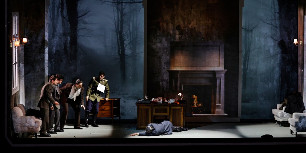 A theatrical scene showing multiple actors in formal attire, gathered around a man lying on the floor in a living room setting, with a backdrop of trees and a fireplace.