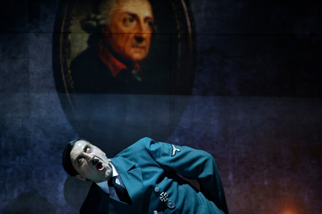 A theatrical scene featuring a man portraying an intense character in a blue military uniform, with a dramatic expression, set against a backdrop displaying a historical portrait.