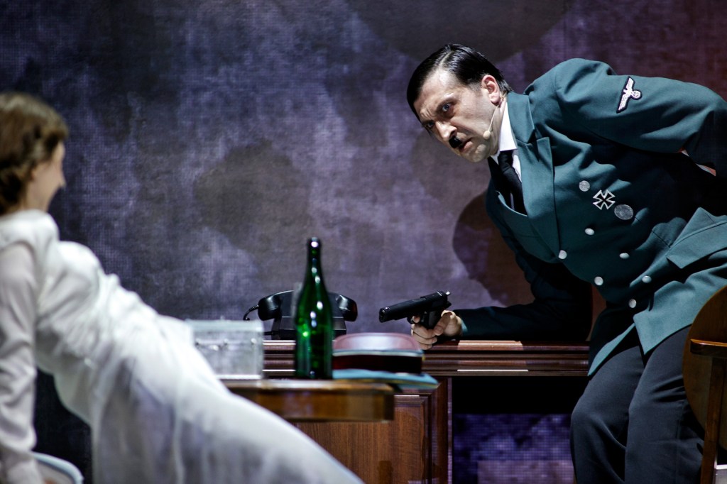 A tense scene depicting a man in a Nazi uniform aiming a gun at a woman seated nearby, set against a dark background with a vintage telephone and a green bottle on a table.