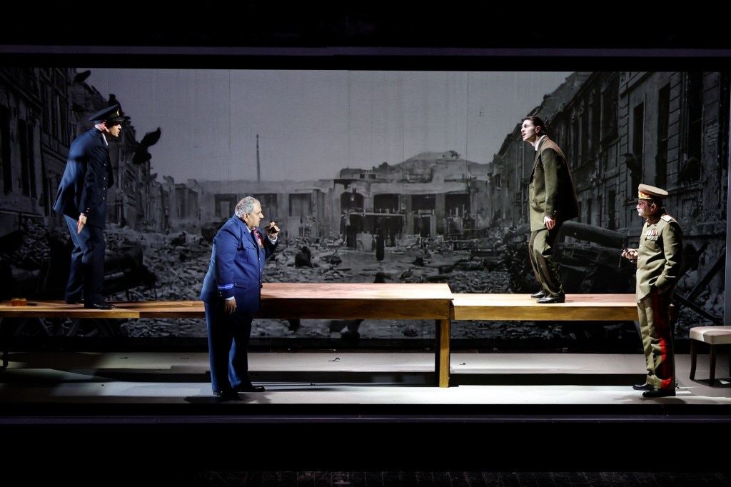 Four actors performing on a stage set with a backdrop of a war-torn city, with one actor on a wooden table speaking into a phone, and others positioned around them.