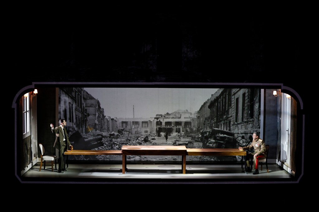 A theatrical scene depicting two characters in a stark, damaged environment, with a large backdrop of war-torn ruins and a table in the center.