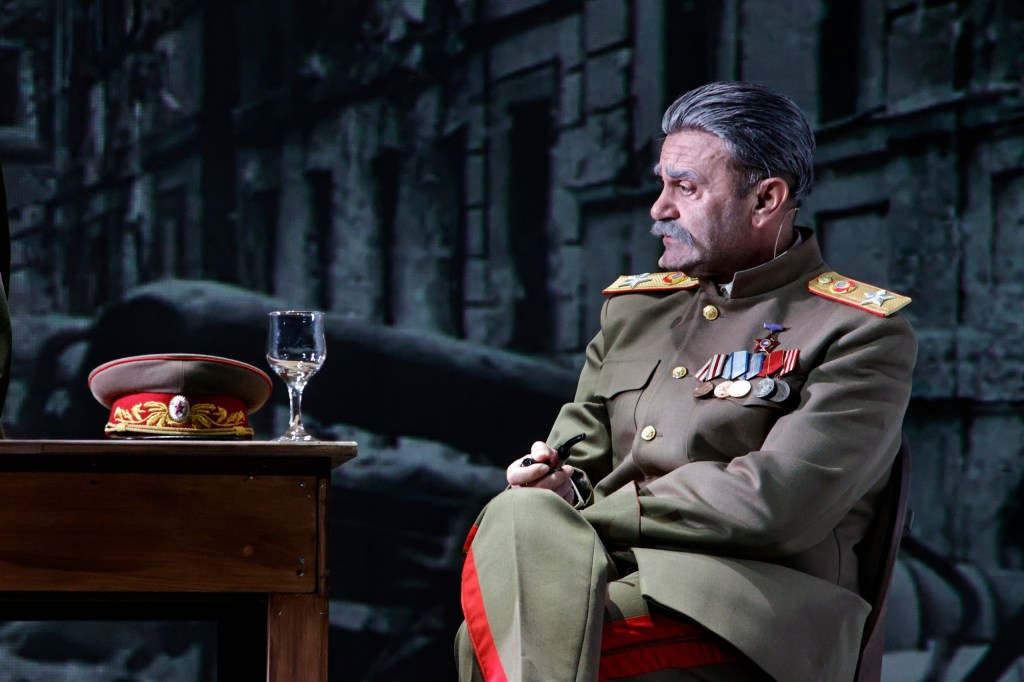 A man dressed in a military uniform with medals sits at a table with a glass and a cap, looking thoughtfully to the side against a blurred background of wartime imagery.