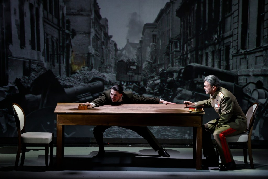 Two men sit at a wooden table during a theatrical performance, with a backdrop depicting a war-torn street. One man leans forward intensely, while the other remains seated, holding a plate.