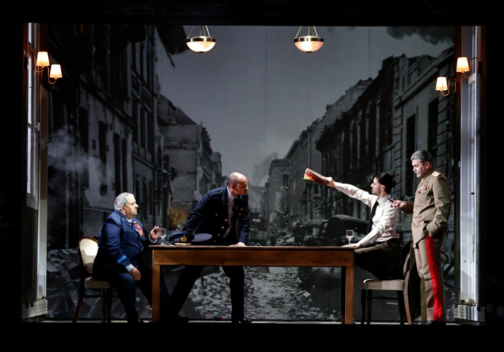 A dramatic scene from a play featuring four characters around a table, with a backdrop of a war-torn city street.