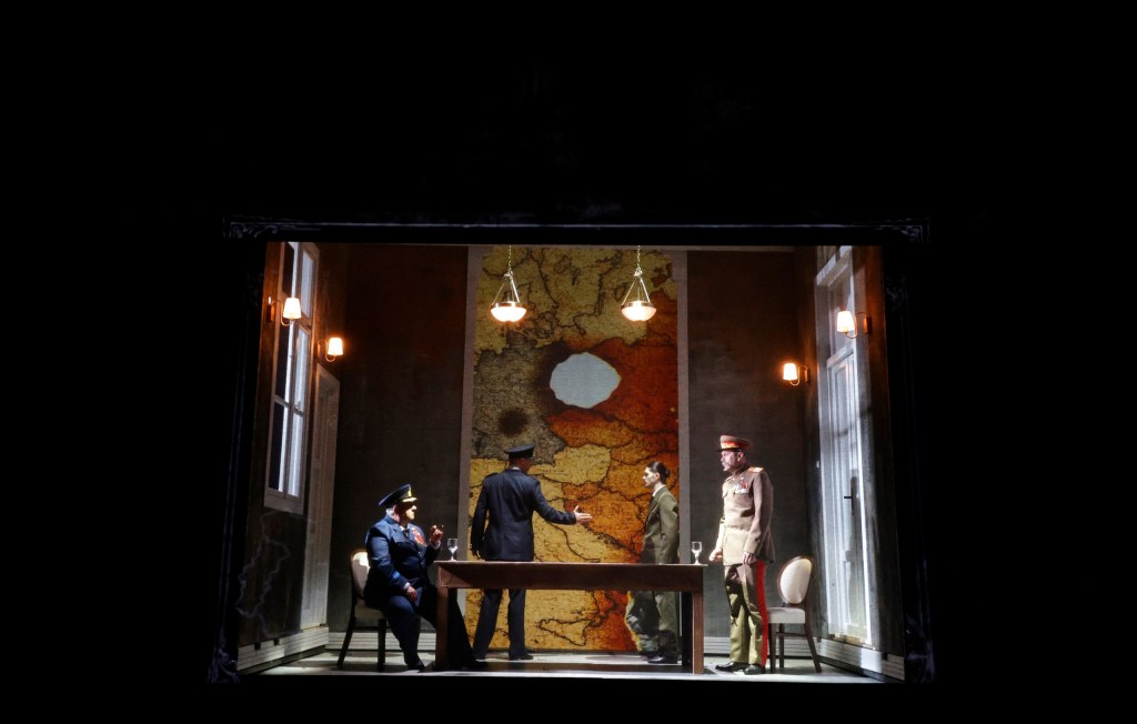 A theatrical scene featuring four male actors in period military attire, engaged in a conversation around a table, with a large map hanging on the wall behind them.
