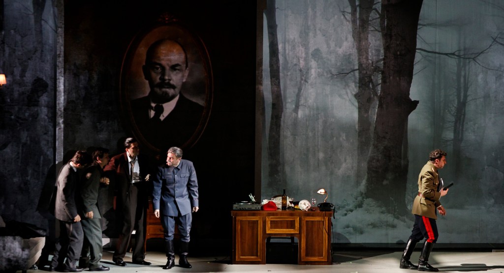 A dramatic scene from a theatrical performance, featuring a group of actors in period costumes interacting in a dimly lit room with a portrait of Lenin in the background.