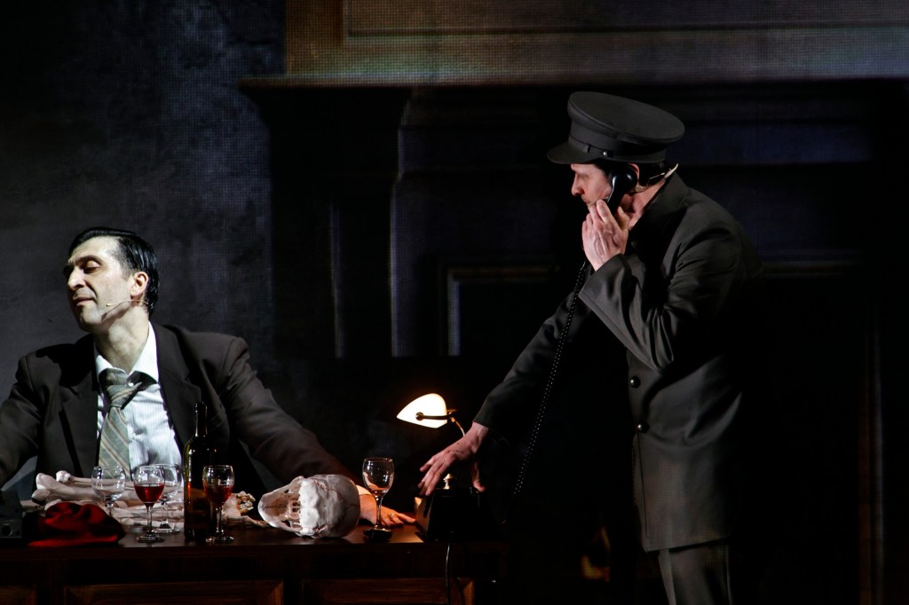 A theatrical scene featuring two actors. One actor, dressed in a suit, is seated at a desk with drinks and a skull, appearing relaxed. The other actor, in a uniform hat, is standing and speaking on a vintage telephone.