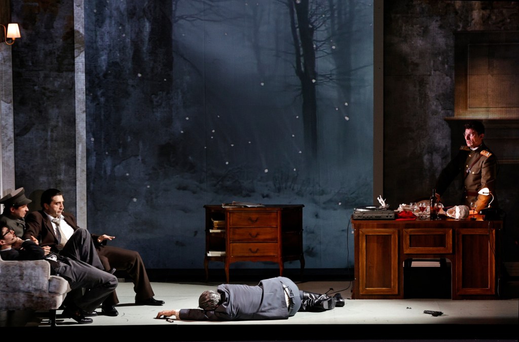 A theatrical scene depicting three men sitting on a couch, with one man lying on the ground, and another man in a military uniform standing at a desk. The backdrop features a snowy forest landscape.