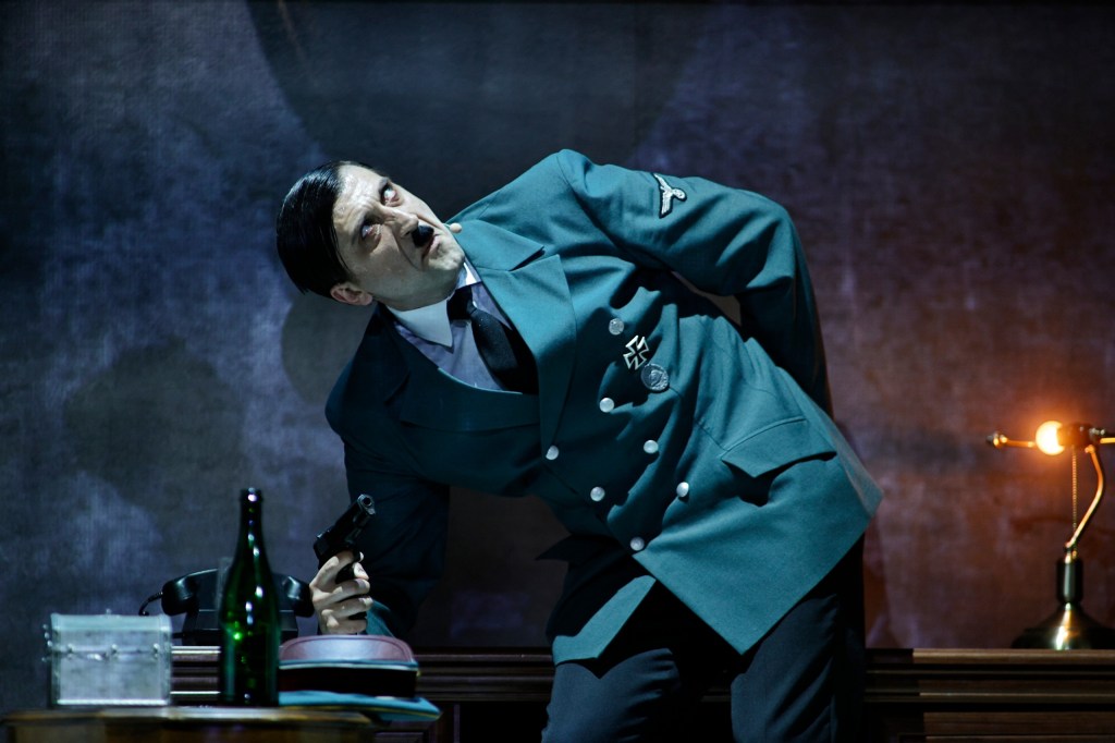 A man in a military uniform holding a pistol, with a tense expression, standing near a desk with a bottle and other items.