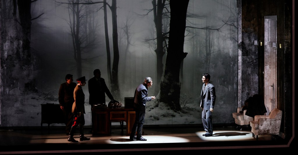 A theatrical scene featuring characters in formal attire on stage, set against a backdrop of dark trees and a wintry landscape.