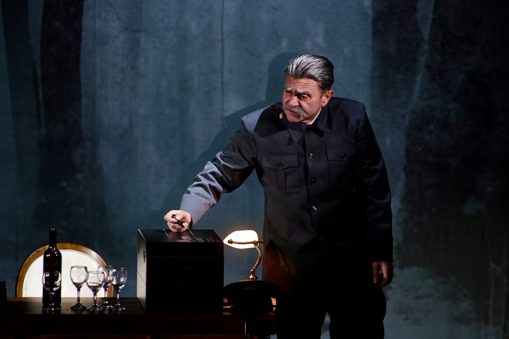 A performer in a dark military uniform stands by a table with a wine bottle and glasses, writing on a box while facing a dramatic backdrop.