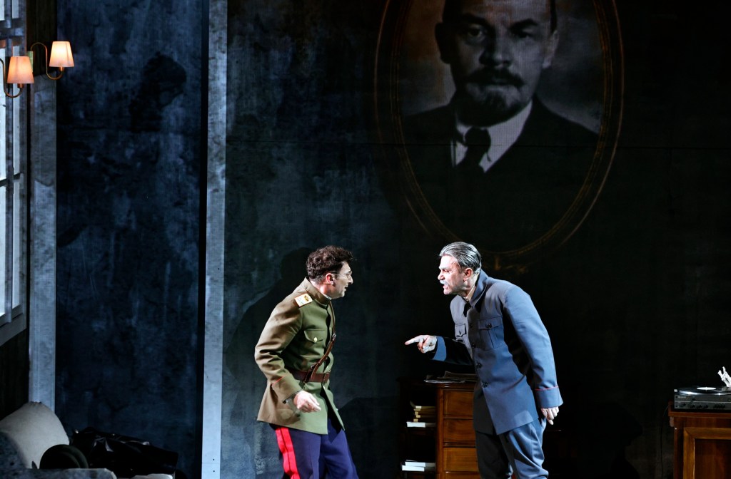 Two actors in a heated confrontation on stage, with one wearing a military uniform and the other in a dark coat. A large portrait of a historical figure hangs in the background.