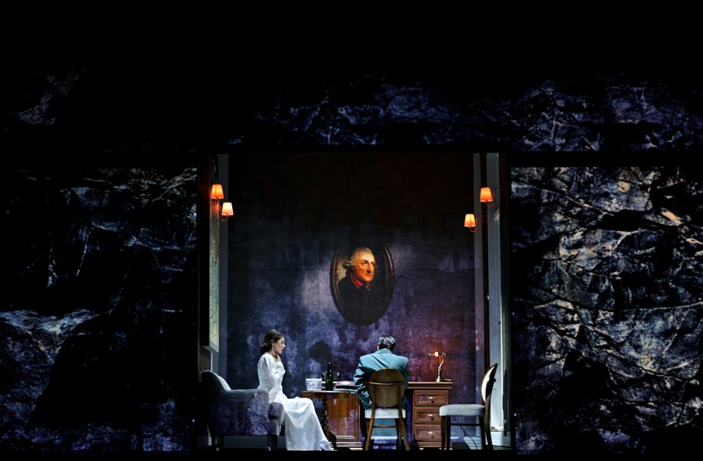 A theatrical scene featuring a woman in a white dress sitting at a table, while a man in a suit sits with his back to the viewer, illuminated by warm light fixtures. A prominent portrait hangs on the wall behind them.