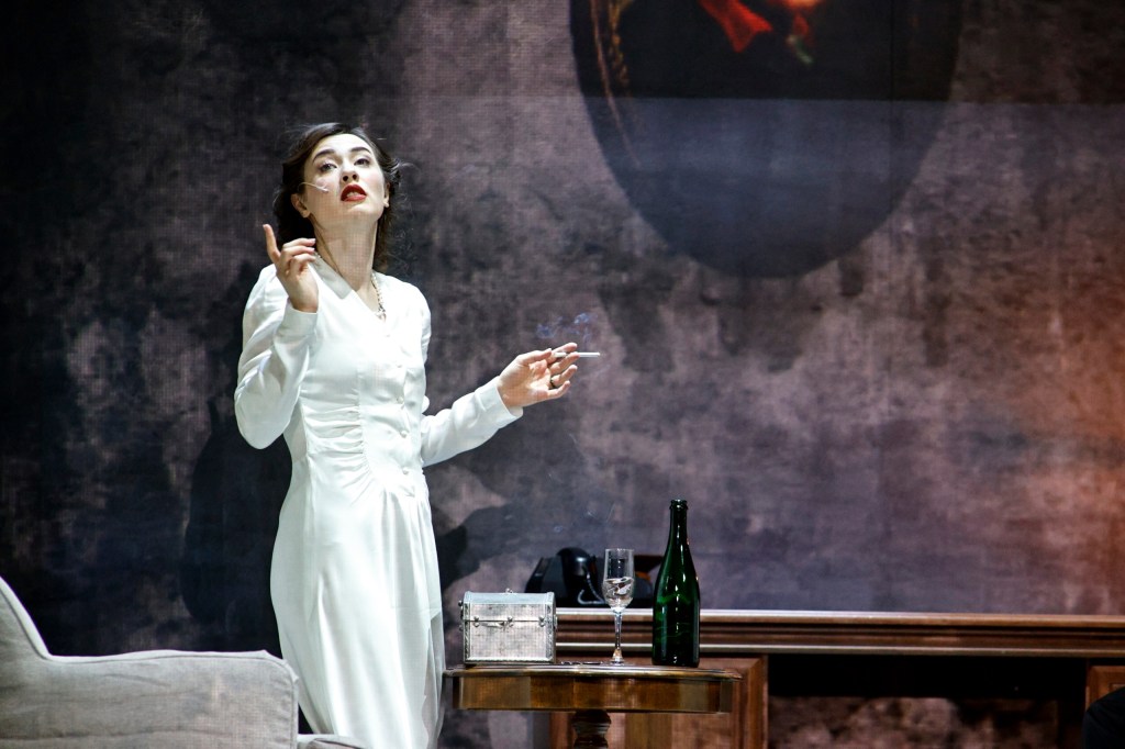 A woman in a vintage white dress stands on stage, gesturing expressively while holding a cigarette. A bottle of champagne and glass are on the table beside her, with an ornate backdrop featuring a painting.