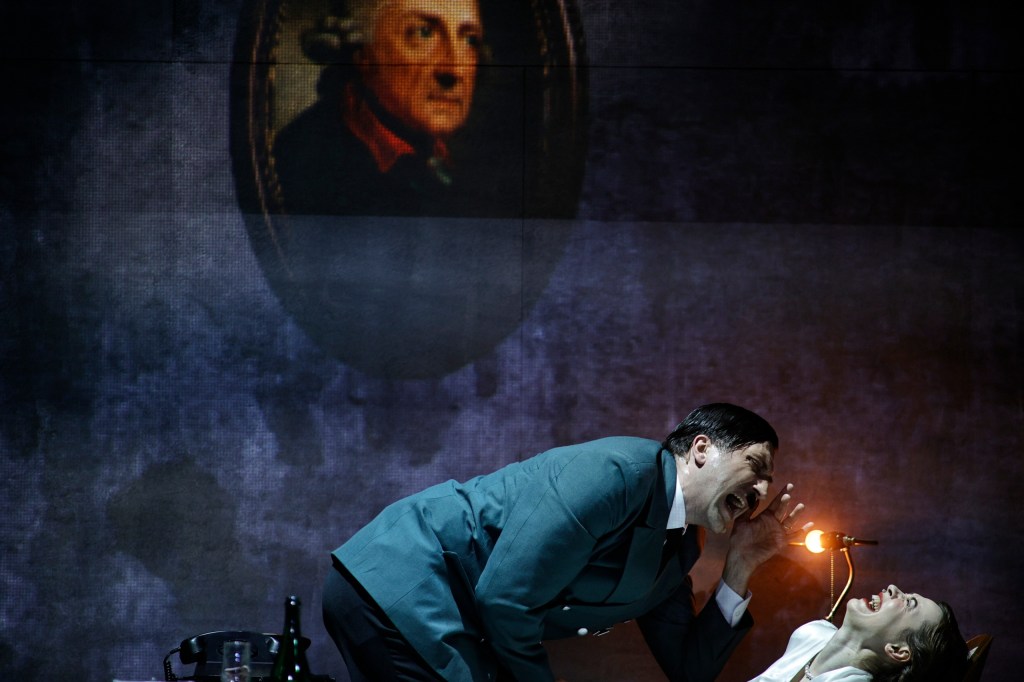 A dramatic theatrical scene featuring a man in a military uniform shouting while kneeling over a woman, who is laughing. In the background, a portrait of a historical figure is projected on a dark stage.
