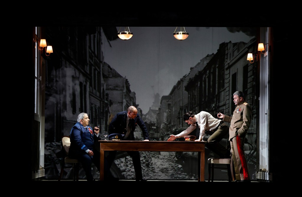 A dramatic scene from a play featuring four characters in military attire around a table, with a backdrop depicting a war-torn city.
