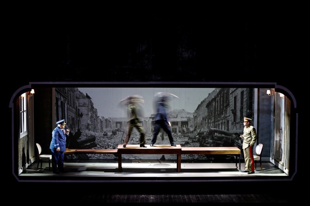 A theatrical scene depicting a dramatic confrontation with two blurred figures on a table and two uniformed men observing from the side, set against a backdrop of a ruined street.