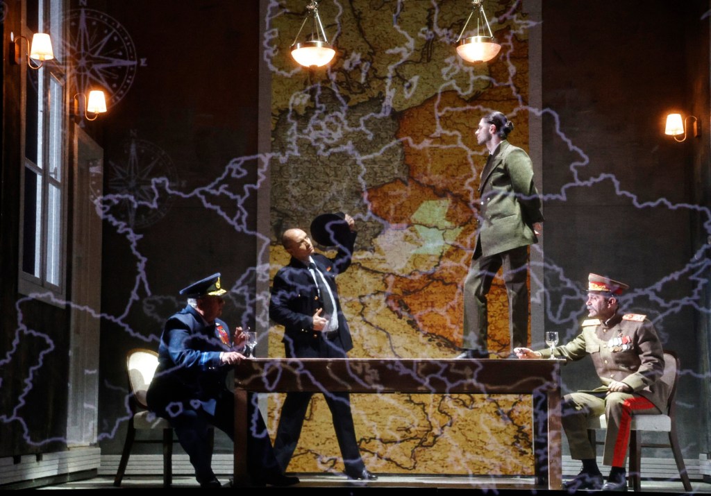 A dramatic scene set in a dimly lit room with a large map projection on the wall. Four characters in military uniforms engage around a table, with one standing assertively while others sit and interact.