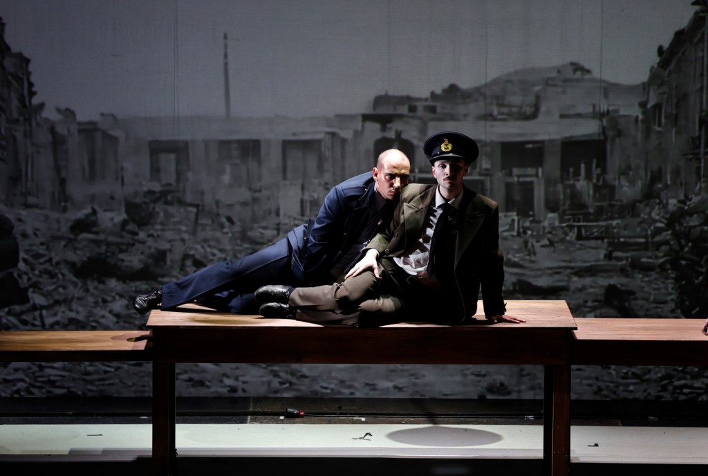 Two actors performing a dramatic scene on a wooden table, set against a backdrop of a war-torn landscape.