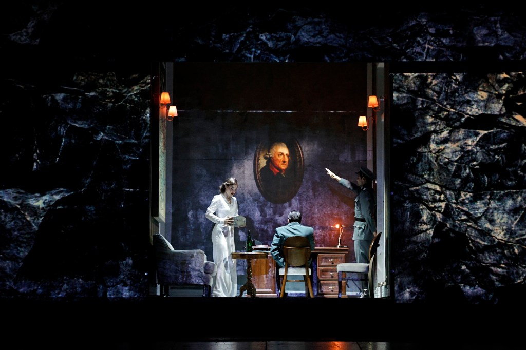 A scene from a theatrical performance featuring a woman in a white dress holding a box, a man seated at a desk, and a uniformed figure pointing, with a portrait on the wall behind them.