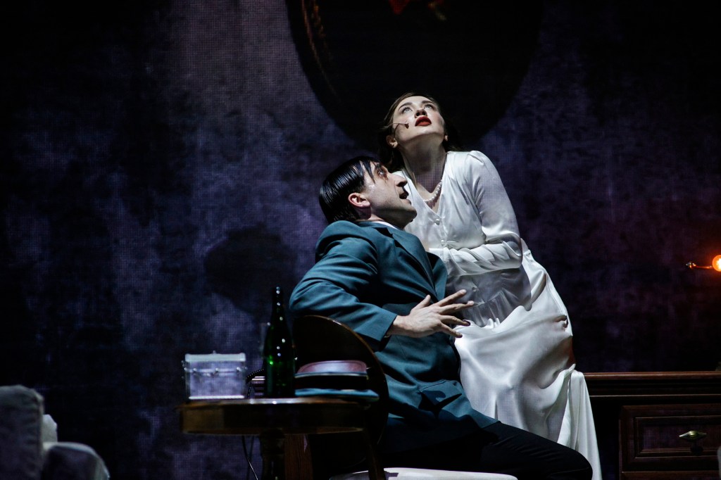 A dramatic scene from a theatrical performance featuring a man in a green suit and a woman in a white dress, both expressing intense emotions. The man is seated, appearing distressed, while the woman stands behind him, looking upwards.