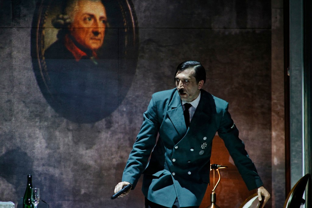 A performer in a military uniform, resembling a historical figure, stands dramatically on stage with a distressed expression, holding a pistol. A framed portrait of another historical figure is projected in the background.