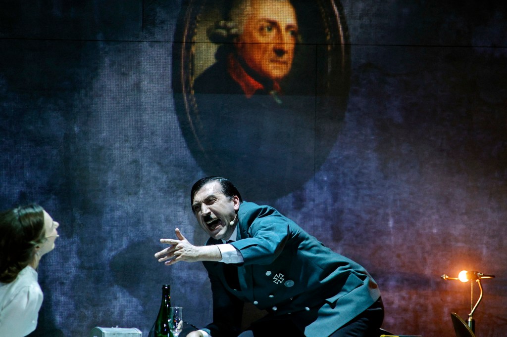 A dramatic theatrical scene featuring a man in a military uniform passionately gesturing, with a woman seated nearby. The backdrop displays a portrait of a historical figure, adding to the intensity of the performance.