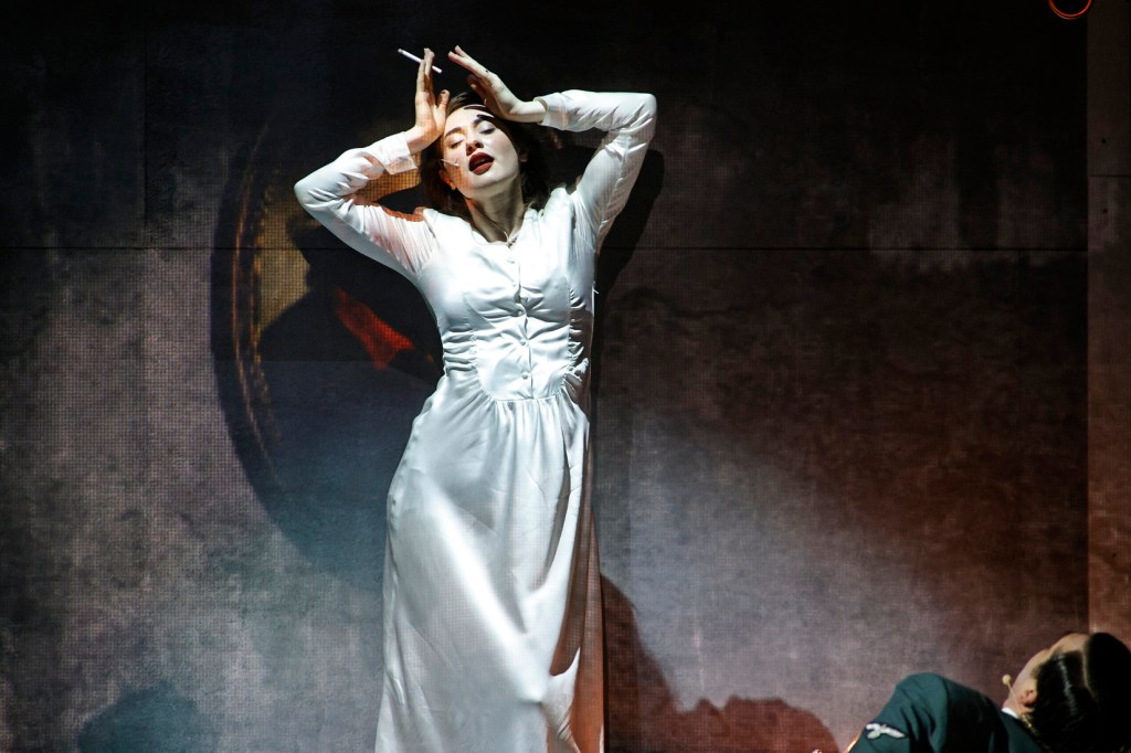 A performer in a white dress striking a dramatic pose, with one hand on her head and holding a cigarette, against a textured background.