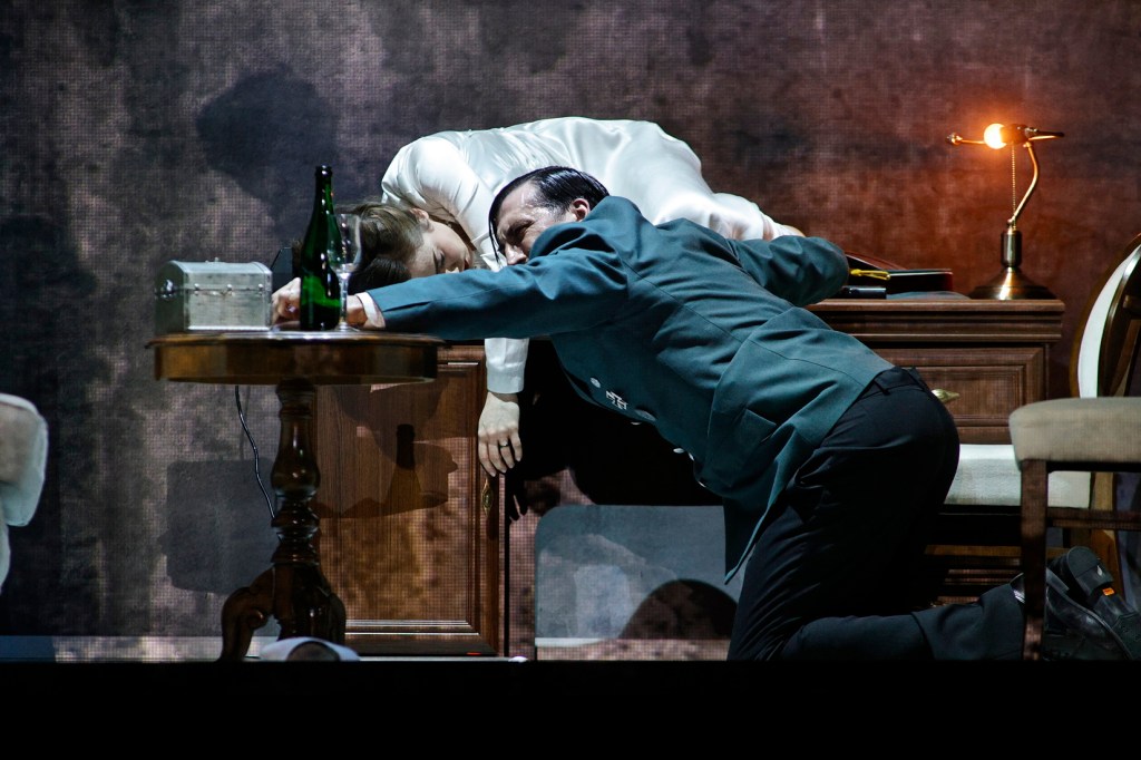 A dramatic scene depicting two performers, one in a military uniform and the other in a white shirt, leaning over a table. A bottle and glass are visible on the table, creating a tense and intimate moment.