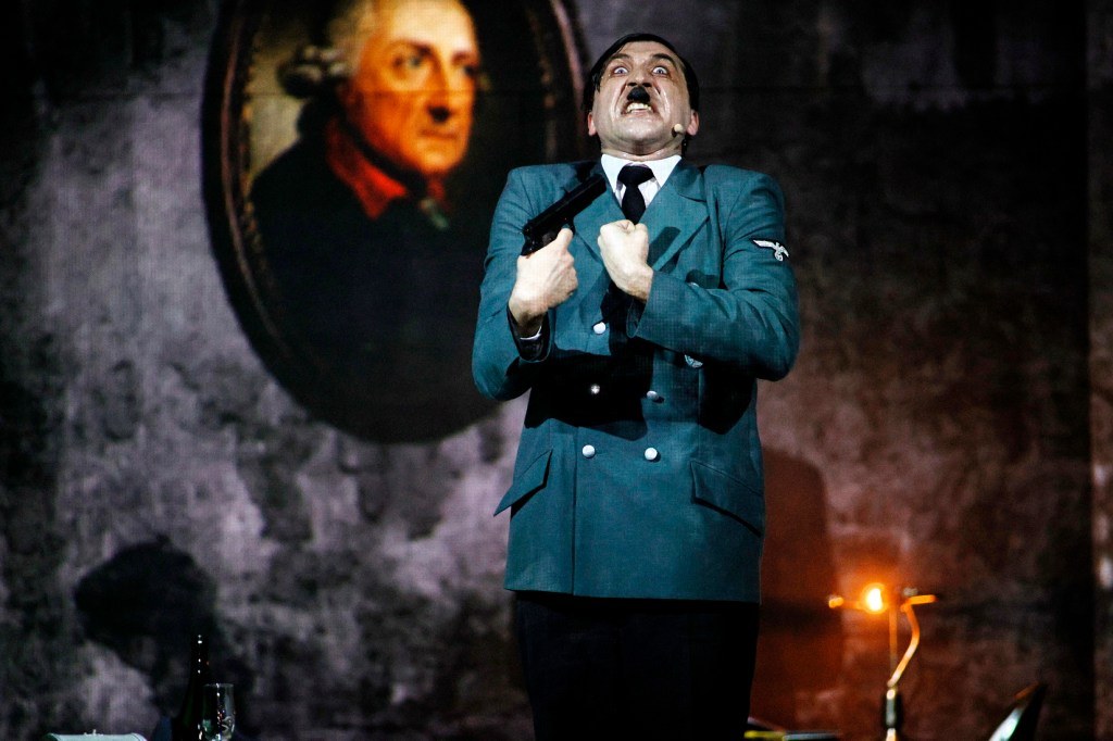 A man in a Nazi uniform dramatically poses with a gun, surrounded by dimly lit props and a portrait in the background.