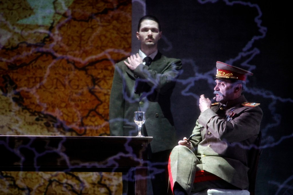A scene from a theatrical performance featuring two men, one dressed in military attire with a hat, sitting at a table with a glass of wine, while a second man stands beside him with a hand on his shoulder. The background displays a projected map with dark, dramatic lighting.
