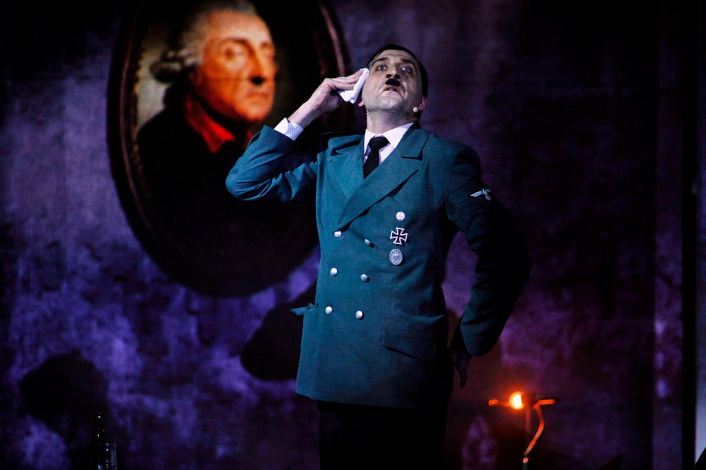 An actor in military uniform performing dramatically on stage, with a historical painting in the background.