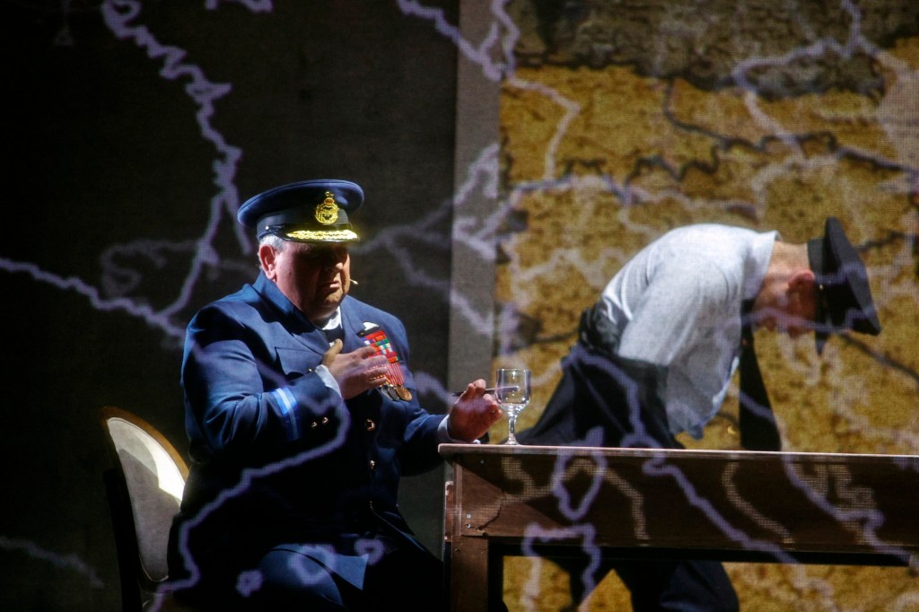 A military officer in a blue uniform and cap sits at a table, holding a glass, while another officer in a gray uniform bends forward, with a backdrop of a map and lightning-like projections.