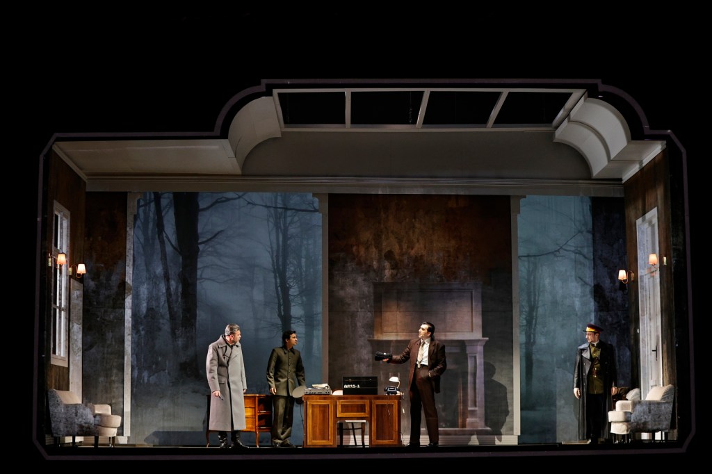 A theatrical scene depicting four characters in a dramatic setting resembling a room with a large window. The backdrop features a dark forest, enhancing the atmosphere. The characters are dressed in period costumes, with various expressions and postures suggestive of a tense interaction.