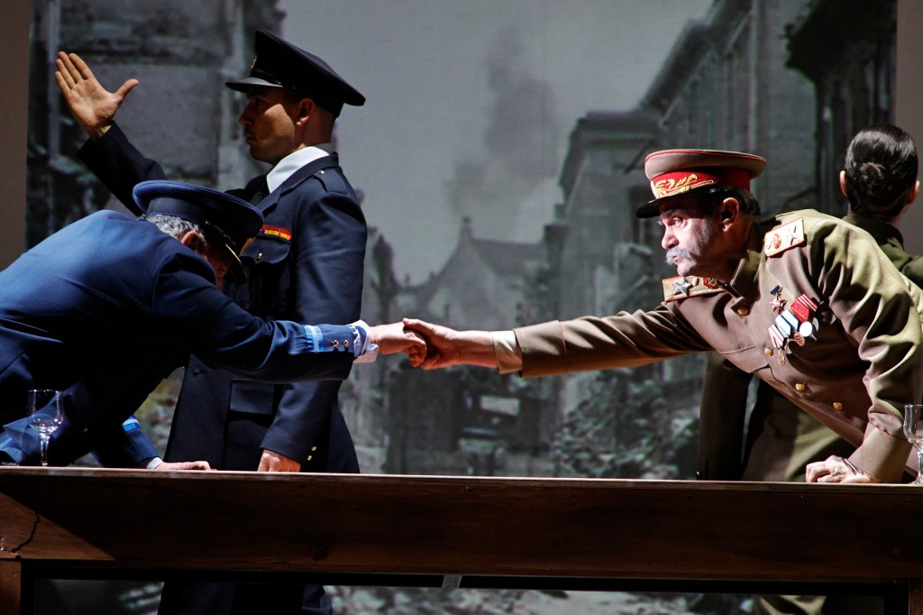 A theatrical scene depicting two military figures shaking hands over a wooden table, with a backdrop of a war-torn city.