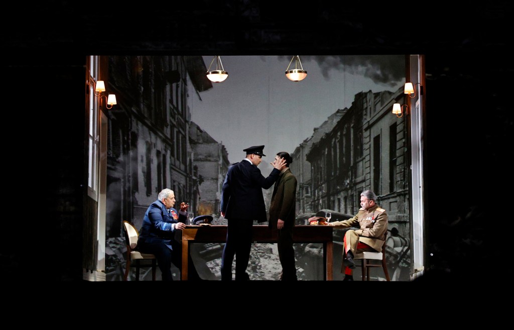 A theatrical scene depicting a tense interaction between a police officer and a man, with two other characters seated at a table in front of a backdrop showing a devastated cityscape.