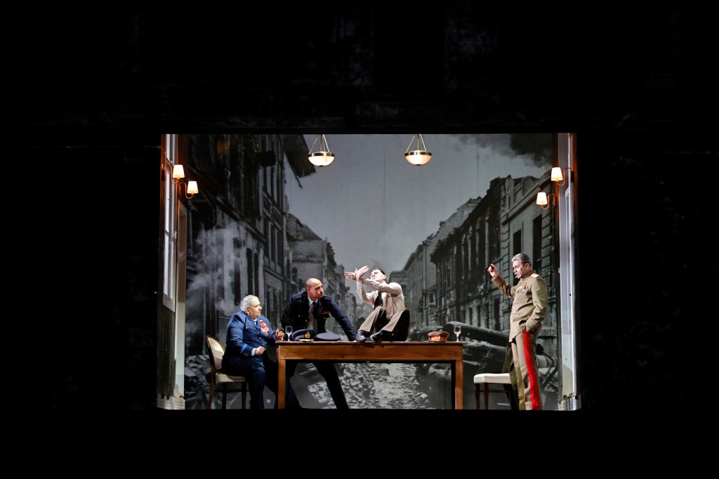 A theatrical scene depicting four characters around a table with a backdrop of a war-torn street.